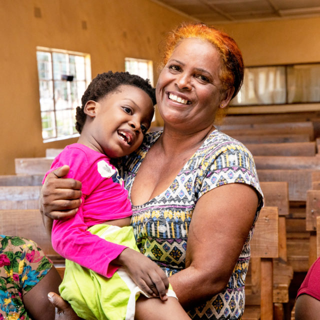 Afrikkalainen nainen hymyilee leveästi kameralle puolilähikuvassa. Hänellä on sylissään lapsi, joka nauraa. Lapsella on kirkkaan pinkki paita ja vihreät housut jalassa. Kuva on otettu sisätilassa, kirkon penkissä.