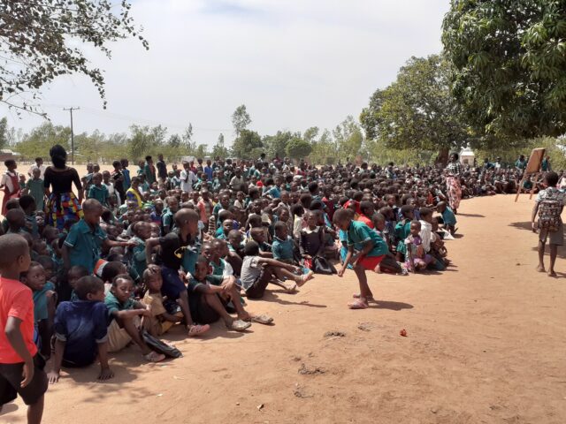 Kuva ulkona hiekkakentällä, taustalla puita. Lapsia ja nuoria afrikkalaisessa kylässä istumassa ja seisomassa tiiviissä ryhmässä katsomassa samaan suuntaan.