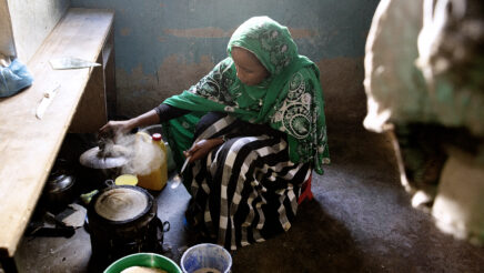 Ylhäältä päin otetussa valokuvassa nainen istuu jakkaralla hiilikeittimen ääressä. Panusta nousee höyryä ja pannulla näkyy taikinasta paistettu ohut lettu. Naisen pään peittona on iso vihreä huivi ja hänellä on pitkä hame. Keittiön lattialla on ämpäreitä ja muita työvälineitä.