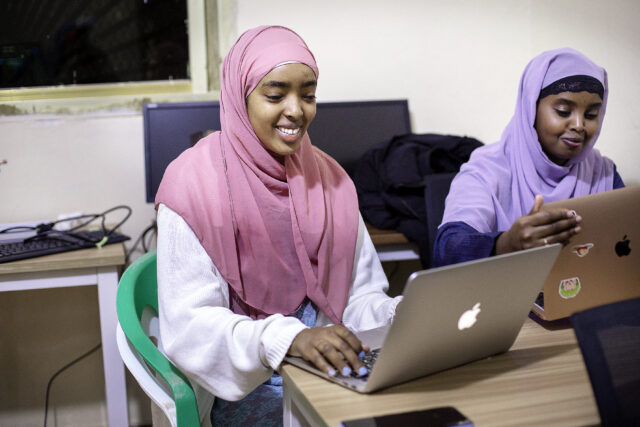 Kaksi nuorta tyttöä istuu työpöydän ääressä kannettavat tietokoneet edessään. Molemmat hymyilevät katsoessaan koneiden näyttöjä. Kummallakin on pään peittävä huivi, yhdellä vaalenapunainen, toisella violetti