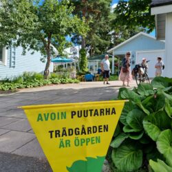 Piha, jossa asfalttia ja sininen talo, sekä banneri jossa lukee avoimet puutarhat.