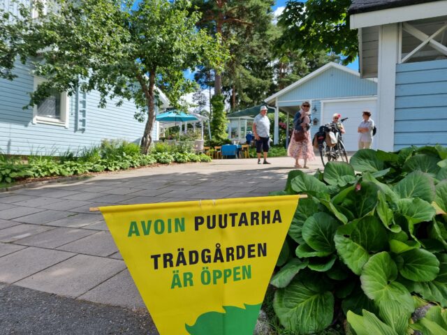 Piha, jossa asfalttia ja sininen talo, sekä banneri jossa lukee avoimet puutarhat.