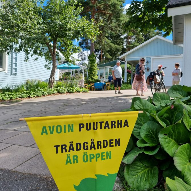 Piha, jossa asfalttia ja sininen talo, sekä banneri jossa lukee avoimet puutarhat.