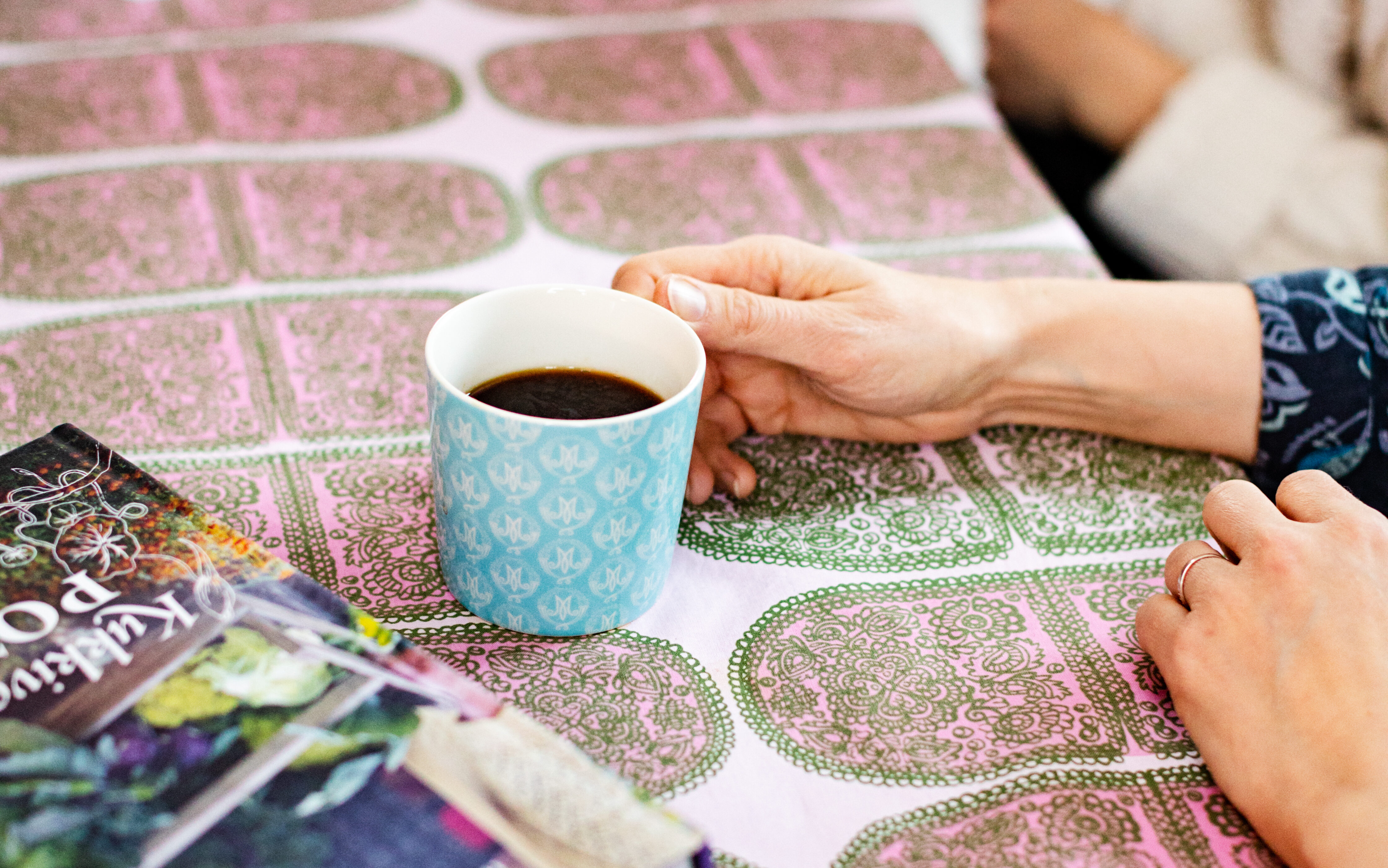 Pöydän päällä on värikäs pöytäliina, kaksi värikästä kirjaa ja kasa paperia. Pöydän ääressä istuu kaksi ihmistä ja molempien edessä on kahvikuppi.