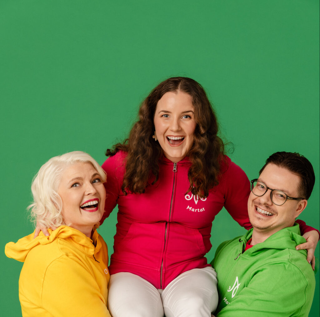 Kuvassa kaksi naista ja yksi mies katsovat iloisesti kameraan. Kuvan reunalla seisovat mies ja nainen kannattelevat käsillään heidän sylissään keskellä istuvaa naista. Kaikilla on päällään värikkäät martta-hupparit.